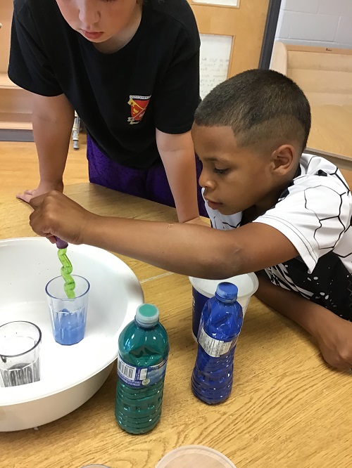 school aged child leaning on table top using dropper to drop blue liquid into cup