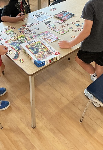 3 school age children standing around a table with a box, some completed puzzles and posters of what each puzzle should look like