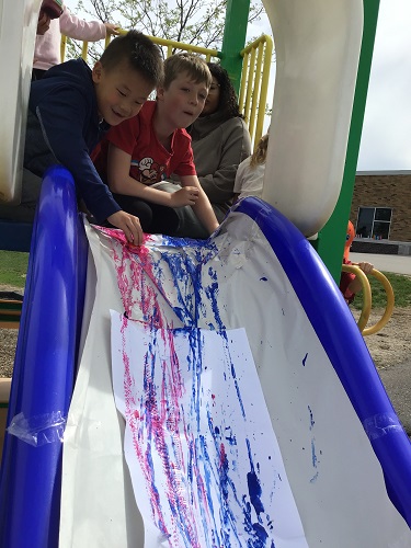 Children at the top of the slide pushing cars through paint and leaving a trail down the slide