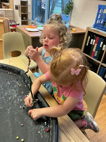 2 preschool children sitting at the table with the tuff tray in front of them, both children are holding a pipe cleaner and attempting to place a bead on the pipe cleaner