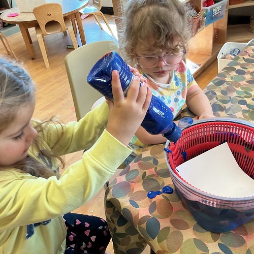 2 preschool children sitting at thet able, one child is holding the bottle of paint upside down with a bowl and paper in front of her, the other child is observing beside