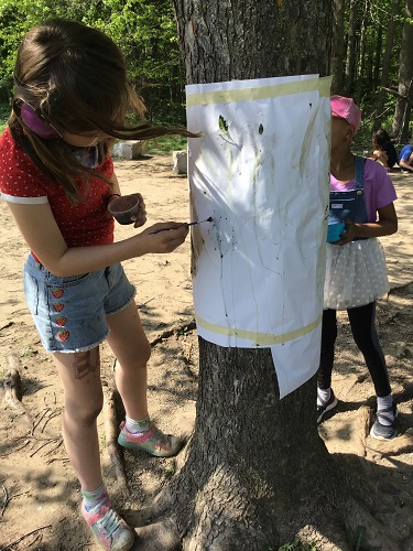 2 children standing by a tree that has white paper taped around it. They have a bowl in one hand and a paint brush in the other - there is drops of paint dripping down the paper taped to the tree trunk
