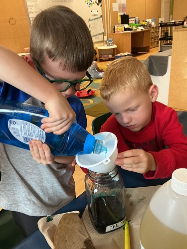 Child pouring dish soap into a jar from a funnel while another child watches