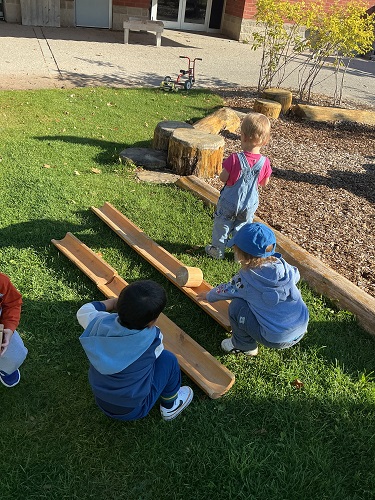 A few children outside rolling pieces of wood down wooden ramps