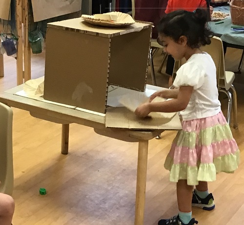 Preschool child holding her pizza and putting into the cardboard box that is the pizza oven