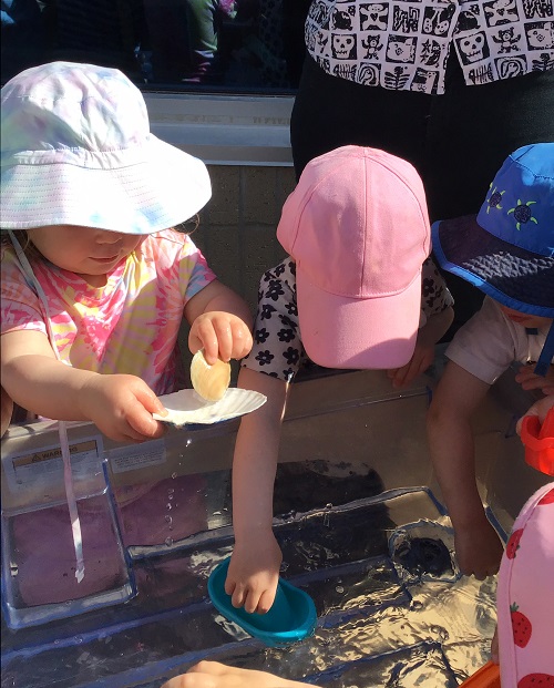 3 toddler children standing at water bin picking up sea shells